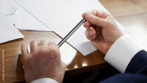 Fotografie Elderly man sits at a table with white sheets of paper and holds a metal pen in his wrinkled hands