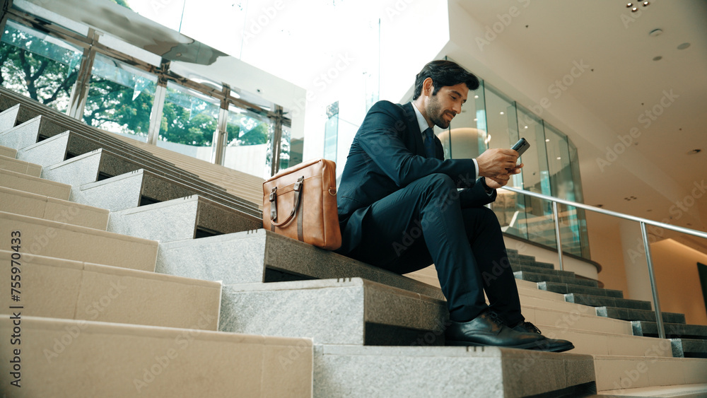Smart project manager looking at mobile phone while sitting at stairs. Attractive caucasian business man working by using phone. Professional investor using phone to plan business strategy. Exultant. - obrazy, fototapety, plakaty 