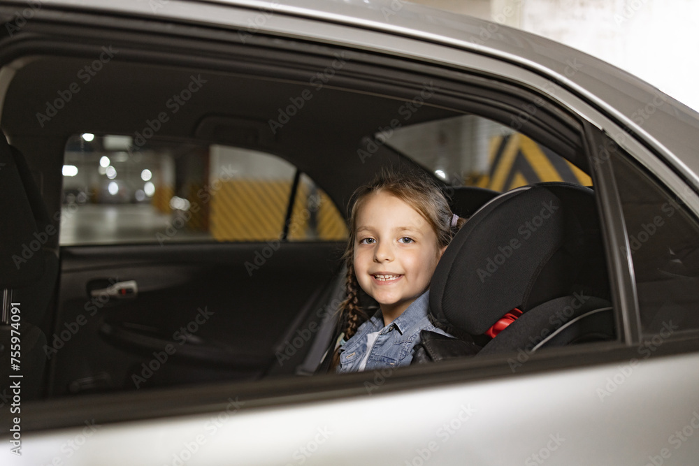 Caucasian little girl sitting on back seat of modern car in child chair and looking at camera from open window against background of underground parking lot. Caucasian little girl sitting on back seat of modern car in child chair and looking at camera from open window against background of underground parking lot.