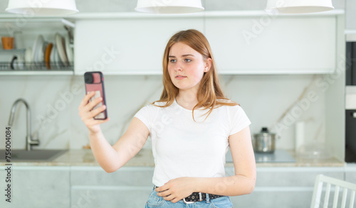 Portrait of European girl teenager in casual clothes using mobile phone and taking pictures of kitchen