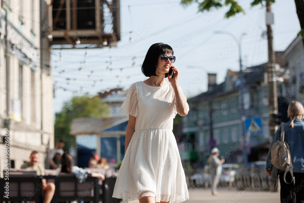 Romantic young brunette woman with short hair wears mini white dress and sunglasses, talking on cellphone, walking on a crowded city street in the summertime. Girl communicates by smartphone outdoors