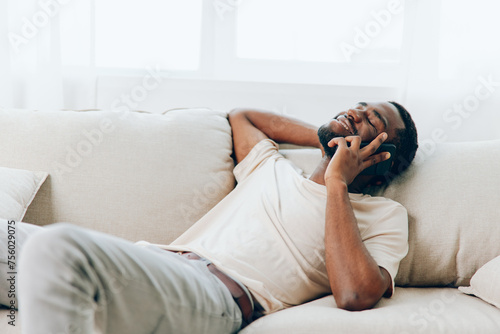 Happy African American man sitting on a black sofa, casually chatting on his smartphone while enjoying a moment of relaxation at home With a confident smile on his face, he effortlessly connects with
