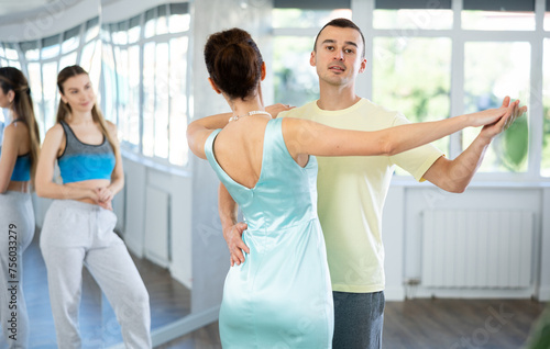 In spacious choreography studio, positive young adult man practicing elegant Viennese waltz with slender brunette in silk aquamarine dress during dance class © JackF