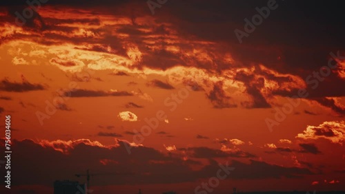 The sky is ablaze with deep red clouds at sunset, casting dramatic shadows over a city silhouette with visible construction cranes. photo