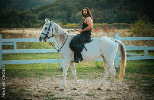 Man equestrian on his horse riding at nature. Attractive man sitting on white horse on the ranch in autumn. Full length of young handsome man sitting on his stallion at the country side.