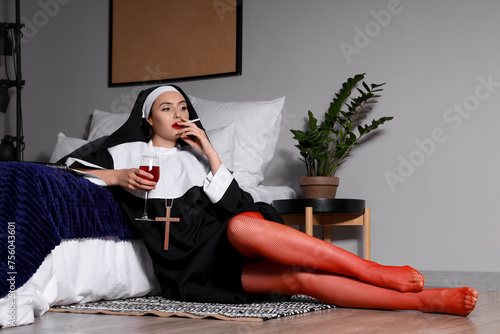 Naughty nun with glass of wine smoking cigarette in bedroom