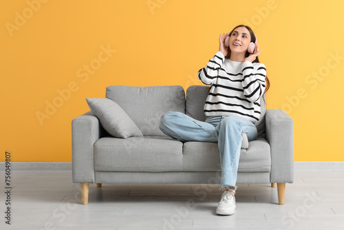 Beautiful young happy woman in headphones sitting on comfortable sofa and listening music near yellow wall