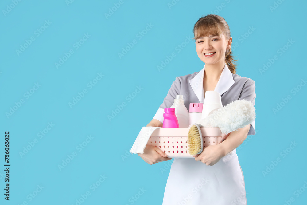 Young chambermaid with cleaning supplies on blue background