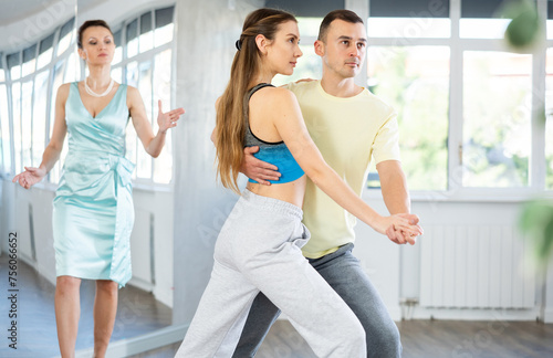 Positive young adult woman and man in sportswear confidently performing tango steps at dance rehearsal in choreography studio while female teacher observing from background © JackF