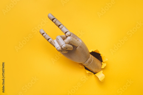 A wooden hand protrudes from a torn hole in yellow paper and shows two fingers, index and little finger, horns (rock) gesture. Concept of love, approval and admiration. Artificial intelligence.