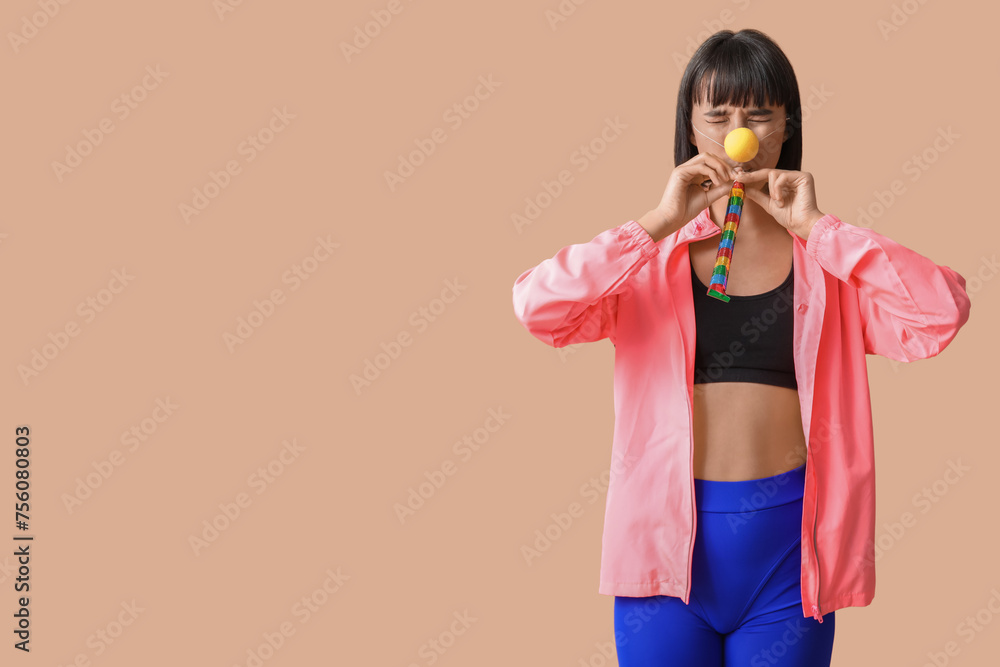 Funny young female fitness trainer with party blower on beige background. Fool's day Funny young female fitness trainer with party blower on beige background. Fool's day