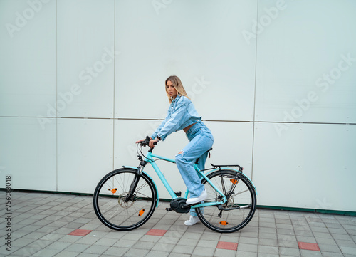 Attractive young blonde dressed in denim on modern blue electric bicycle, posing 