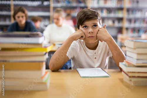Portrait of a thoughtful ten-year-old boy sitting at a desk in the school library among textbooks