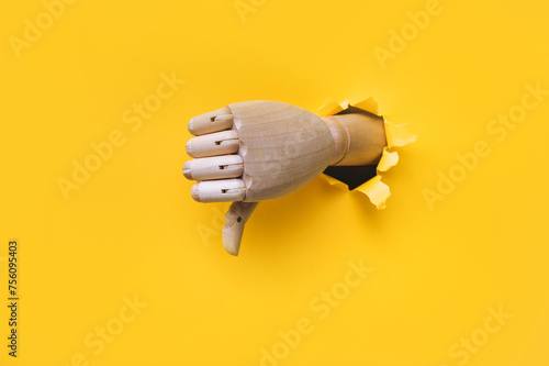 A right wooden hand protrudes from a torn hole in yellow paper and shows a thumbs down (dislike). The concept of disapproval and that things will go badly. Artificial intelligence, robot.