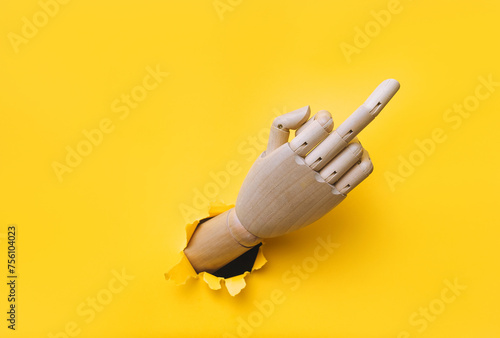 Middle finger of a rsght wooden hand, offensive gesture. Torn hole in yellow paper. Fuck you concept. Aggressive reaction of artificial intelligence or robot. photo