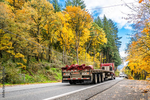 Industrial red big rig semi truck transporting empty flat bed semi trailer with rolled up tarp on the back running on the autumn narrow road with yellow trees on the hills