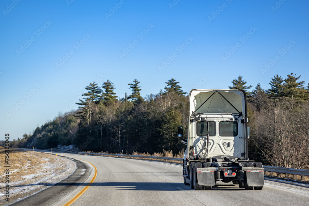 White big rig semi truck tractor without semi trailer running on the one way winter highway road with trees on the side