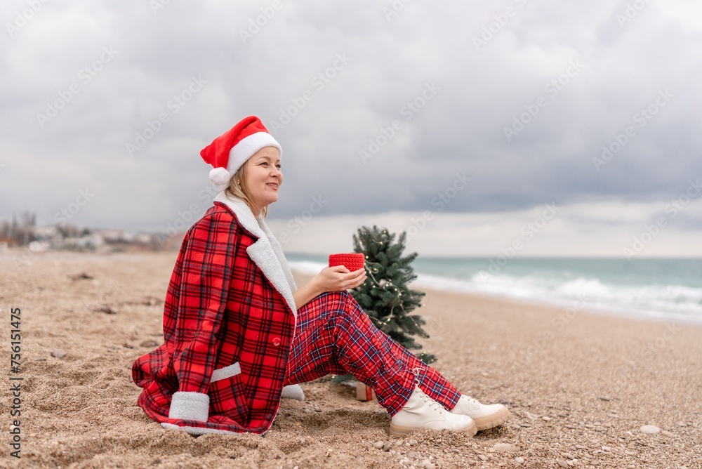 Obraz premium Sea Lady in Santa hat plaid shirt with a red mug in her hands enjoys beach with Christmas tree. Coastal area. Christmas, New Year holidays concep