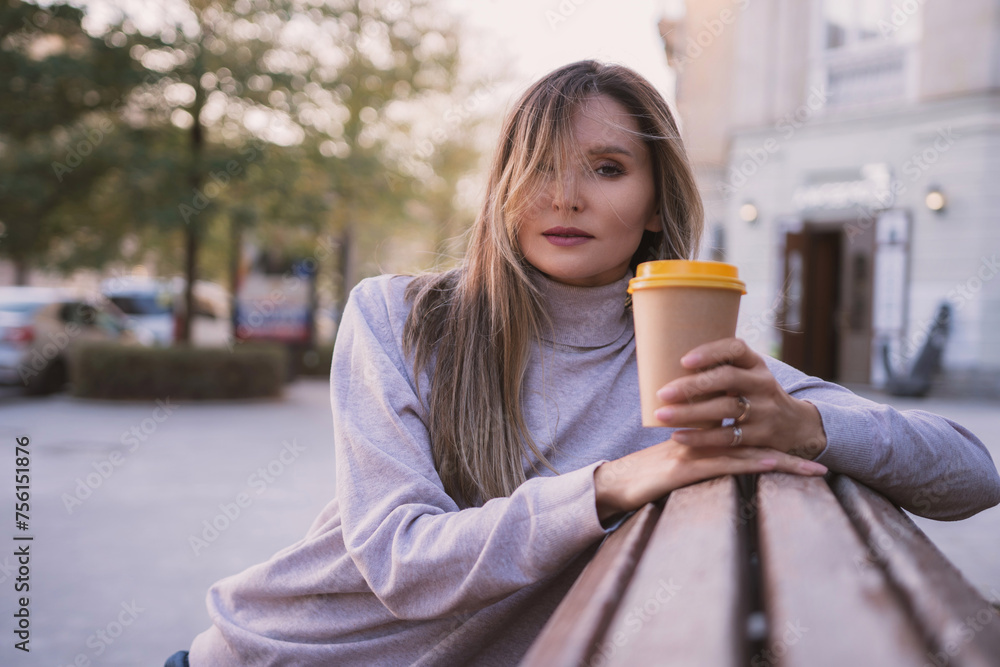 Woman drinks from cup on wooden bench. She is wearing a white shirt enjoying her beverage. The bench is located in a park setting, with trees in the background. Woman drinks from cup on wooden bench. She is wearing a white shirt enjoying her beverage. The bench is located in a park setting, with trees in the background.