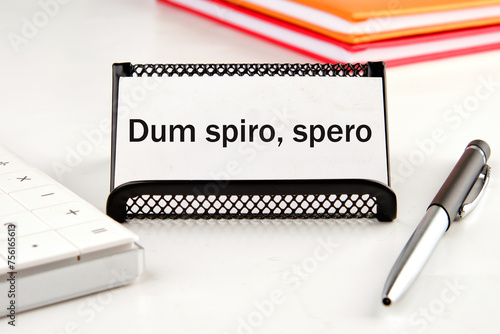 Dum Spiro Spero - latin phrase means While I Breath, I Hope. on a white business card next to a calculator, notepad and pen photo