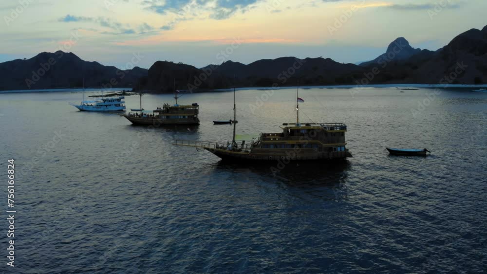 Traditional Wooden Tour Boats anchored off the coast of Padar Island, near Komodo in Indonesia. Drone shot during the evening with mountainous island and night sky In the background. Traditional Wooden Tour Boats anchored off the coast of Padar Island, near Komodo in Indonesia. Drone shot during the evening with mountainous island and night sky In the background.