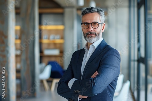 Mature male executive with a beard and glasses standing arms crossed in a contemporary office setting.