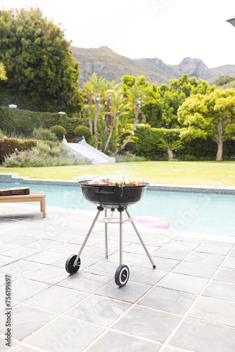 A charcoal grill is ready with food on it, beside a pool in a lush garden with copy space