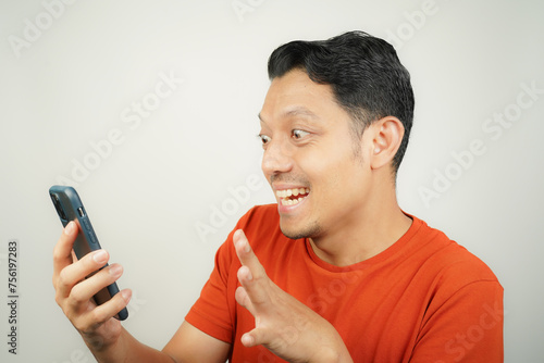 Asian man in orange t-shirt happy looking at what's on smartphone on isolated background © Aria Armoko