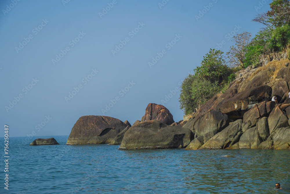 Man wearing sun glass enjoying and swimming into the sea near shore. Big rocks and green forest background on an island. Butterfly beach in Goa, India. Blue ocean water with huge rocks and nearby hill