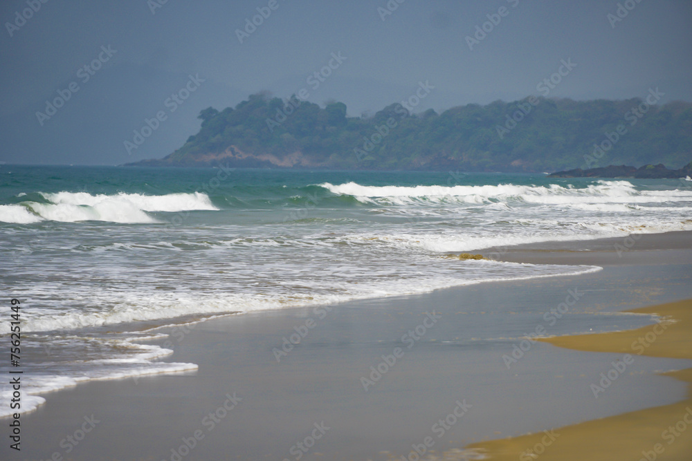 Beautiful sandy beach in Goa, India. Low tides in Sea on a sunny day. Green forest hills near sea shore. Brown sandy beach and white waves in water. Sun reflection in sand when water hits the sand.