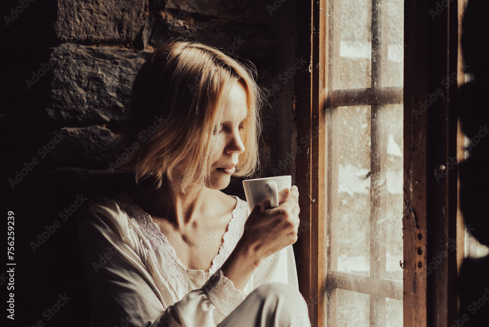 Obraz premium A woman with a pensive look drinks coffee at the window. Middle age woman in a white shirt. Sad look, thoughts.