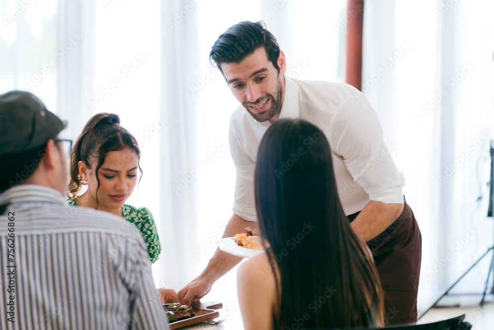 Fototapeta premium Waitress man serving food to group of diverse customer in restaurant, eatery client woman and man having smile and happy with service mind from cafes staff, lunch or dinner time lifestyle with family