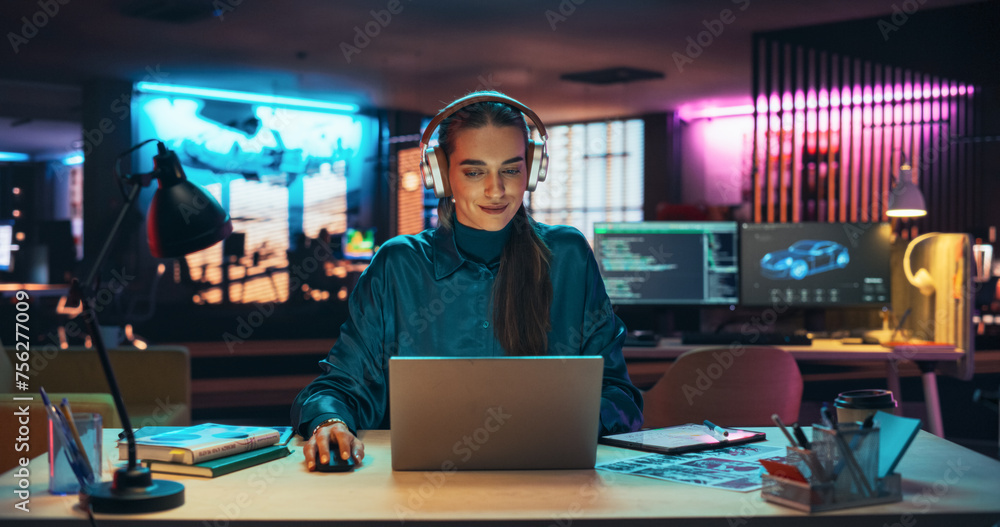 Portrait of a Beautiful Manager Sitting at a Desk in Creative Agency, Using Laptop Computer. Young Stylish Female with Headphones Looking at Camera with Big Smile While Working on New Project Portrait of a Beautiful Manager Sitting at a Desk in Creative Agency, Using Laptop Computer. Young Stylish Female with Headphones Looking at Camera with Big Smile While Working on New Project
