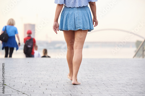 An adult woman in a denim skirt walks barefoot along the city embankment in Kyiv