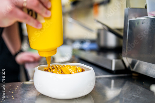 Chef in a professional kitchen drizzling mustard from a yellow bottle onto a dish, with blurred kitchen equipment in the background.