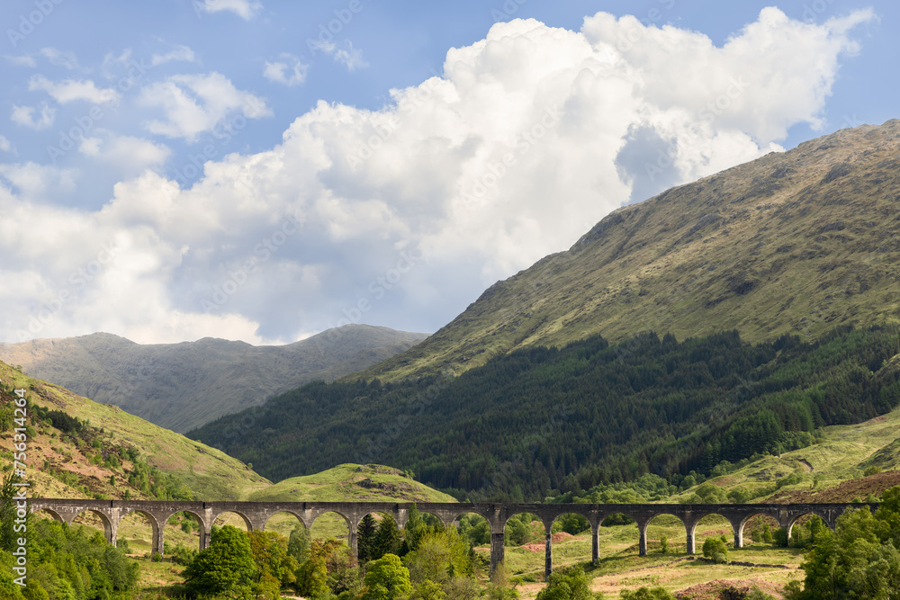 The Glenfinnan Viaduct carves a path through the Scottish Highlands, its arches framing the lush greenery, with mountainous terrain rising under a dramatic sky