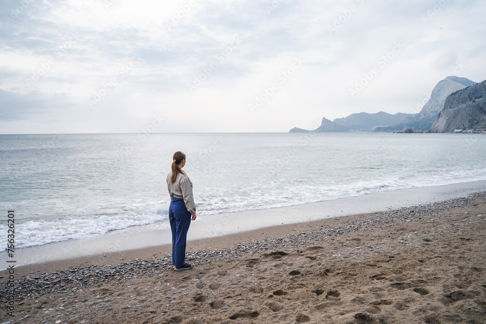 Back side view of a girl stands on the seashore, young woman is looking into the distance, before sunset, dramatic cloudy weather