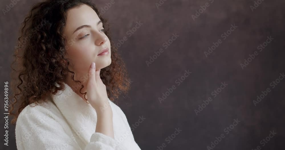 Skin rejuvenation facelift treatment woman face. Young woman in a white bathrobe who gently touches and strokes her face skin from under eyes to the neck and looks at the camera Skin rejuvenation facelift treatment woman face. Young woman in a white bathrobe who gently touches and strokes her face skin from under eyes to the neck and looks at the camera