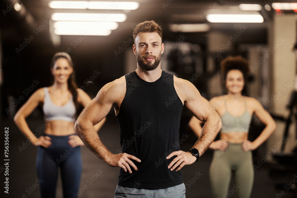 Strong sportsman is standing in a gym and smiling at the camera with sportswomen in a blurry background. Strong sportsman is standing in a gym and smiling at the camera with sportswomen in a blurry background.