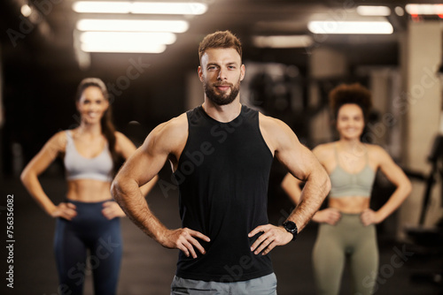 Strong sportsman is standing in a gym and smiling at the camera with sportswomen in a blurry background.