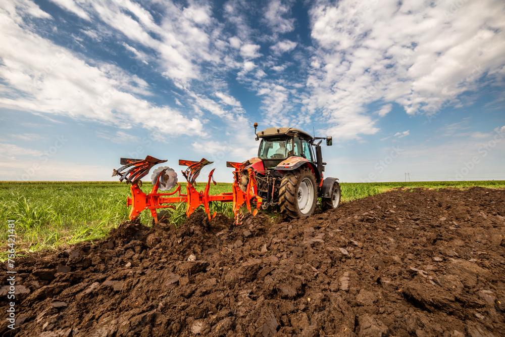 Fototapeta premium Red tractor is engaged in plowing the soil in a vast green farmland under a cloudy sky
