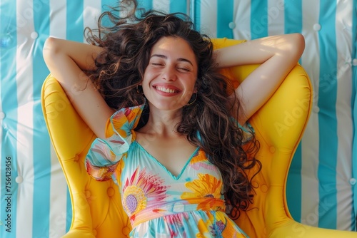 Isolated on a striped blue white background, a pretty fashionable young woman in colorful dress and long curly brunette hair has fun in yellow chair. Summer time, cheerful mood, joy, happiness,