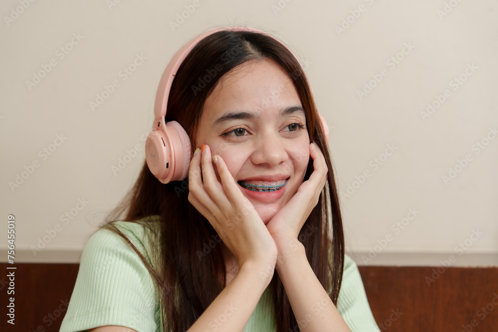 Woman Asian shoulder-length hair wearing light pink headphones, braces gleaming, gazes upward, hand on cheek, lost in music. Female in pastel green top, enjoys tunes, grinning with silver braces Woman Asian shoulder-length hair wearing light pink headphones, braces gleaming, gazes upward, hand on cheek, lost in music. Female in pastel green top, enjoys tunes, grinning with silver braces