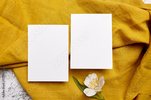 Two white blank greeting cards arranged on a mustard yellow textured linen with a single alstroemeria flower, top view, flat lay