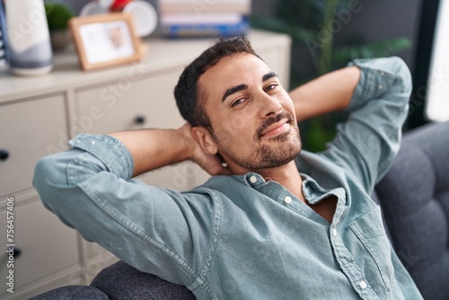 Young hispanic man relaxed with hands on head sitting on sofa at home