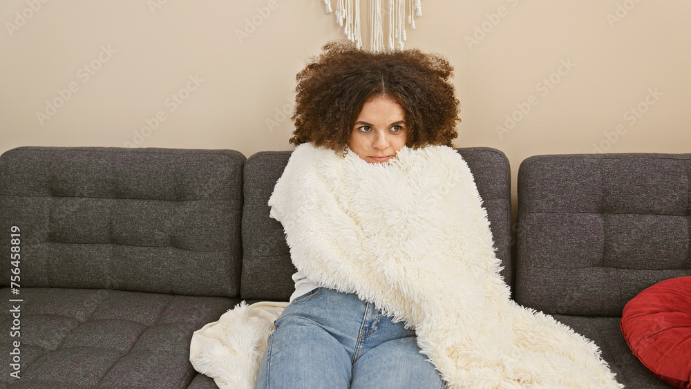 Young hispanic woman with curly hair wrapped in a white blanket sitting on a grey couch indoors. Young hispanic woman with curly hair wrapped in a white blanket sitting on a grey couch indoors.