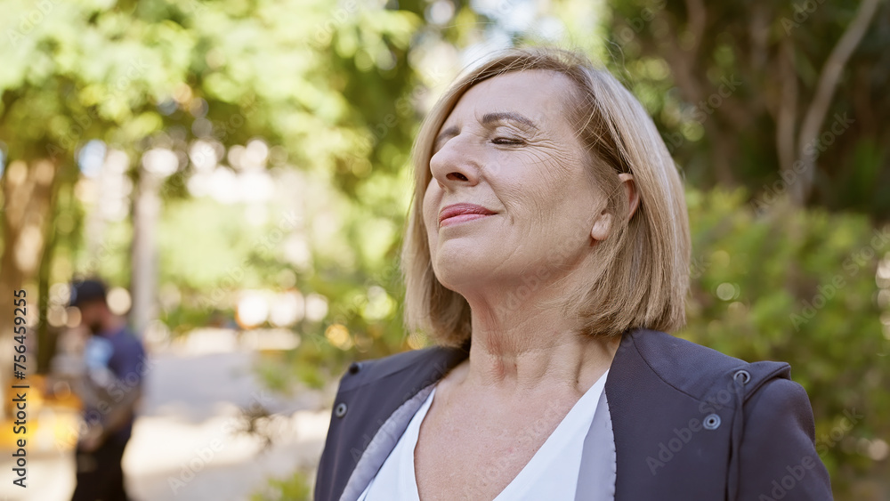 Mature blonde woman smiling contentedly in a sunny park with lush greenery surrounding her