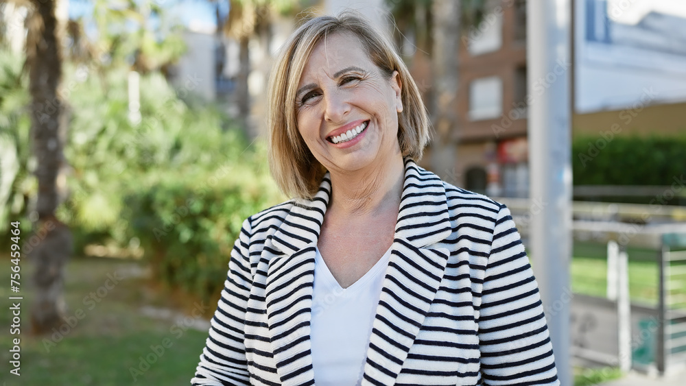 Smiling middle-aged woman outdoors in a striped jacket with greenery and blurred city background Smiling middle-aged woman outdoors in a striped jacket with greenery and blurred city background