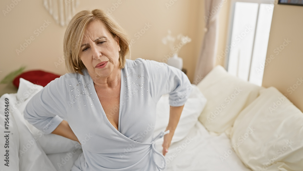 Middle-aged blonde woman in pain holding her back in a bright bedroom at home Middle-aged blonde woman in pain holding her back in a bright bedroom at home