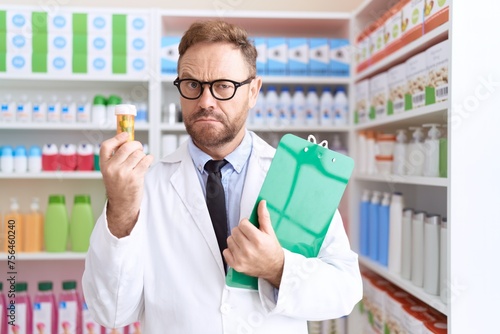 Middle age man with beard working at pharmacy drugstore holding pills clueless and confused expression. doubt concept.